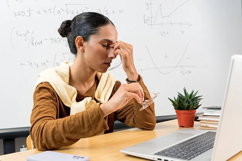 A woman at a desk with her glasses off, pinching the bridge of her nose