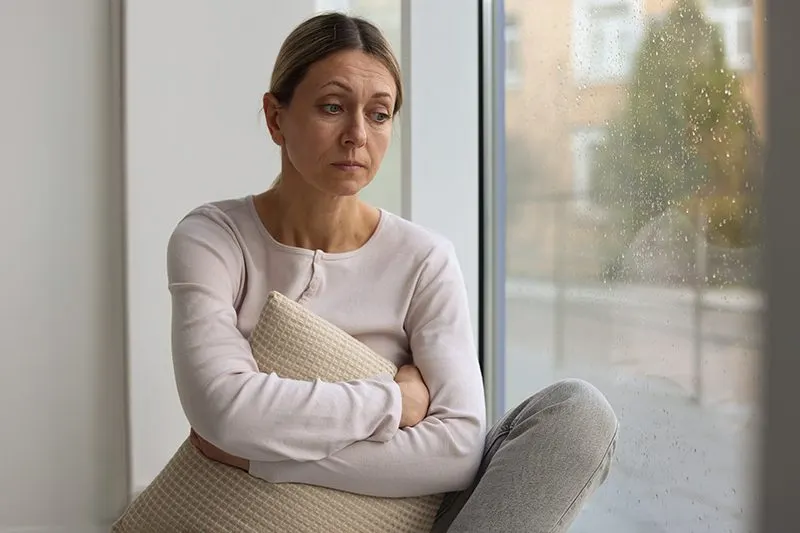 A woman sitting in a window seat holding a cushion, looking away, rain on the glass